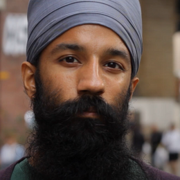 Close up with a person with a long black beard and brown skin and a head covering. 