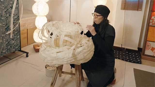 Person wearing glasses and all black kneels in front of stool with lighting fixture on top