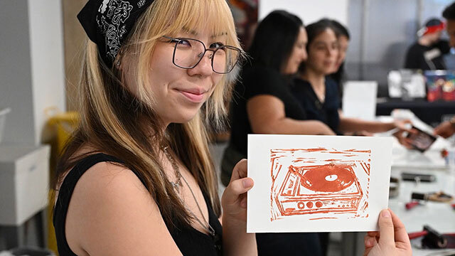 Person wearing bandana smiles, holds linocut print up to camera