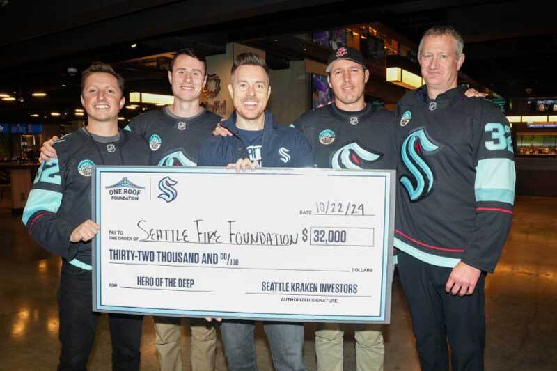 Five Seattle firefighters and rescue swimmers present a giant check for $32,000 provided by the Kraken and One Roof Foundation