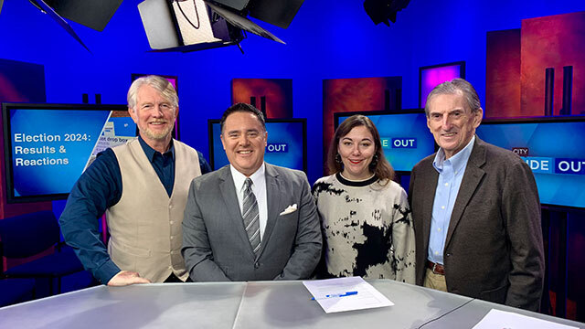 Four people stand in a television studio with blue and red background