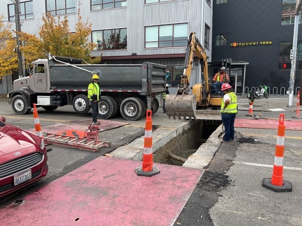 Person runs a backhoe to dig a trench in the road, two other people in construction vests and hard hats stand on either side of the new trench. 