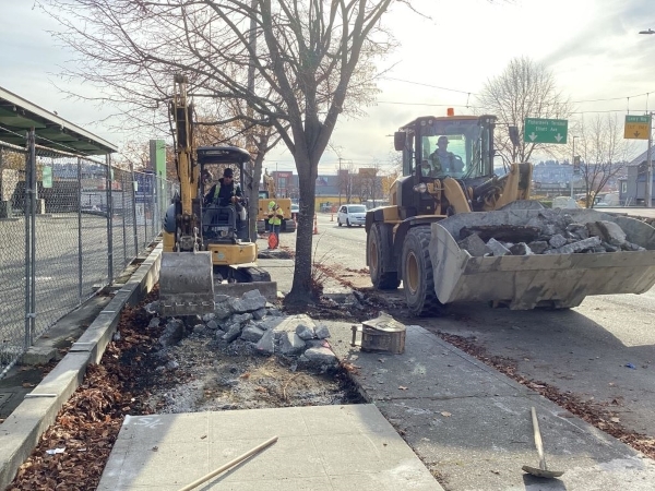 Person runs a backhoe to dig up concrete on an old sidewalk. Another person drives a second backhoe that carries broken concrete away.