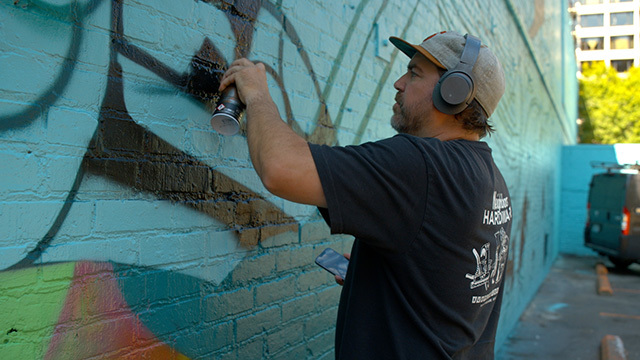 Joe Nix wears headphones, sprays paint on mural on brick wall outdoors