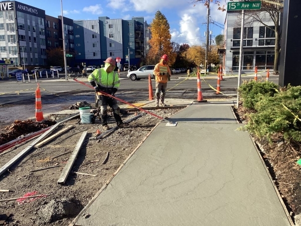 Person in a hard hat and bright yellow jacket uses a long handled tool to smooth newly poured concrete 