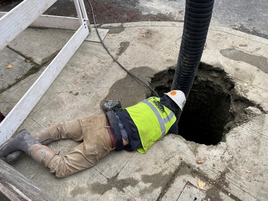 Construction worker laying on the ground looking down at a hole in the concrete for underground utility verification