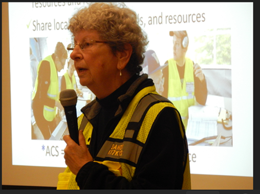 Photo of an older white woman with short curly hair holding a microphone wearing a bright yellow safety vest with a screen behind her