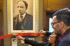 Two people cutting a red ribbon with a black and white portrait hanging in a frame behind it