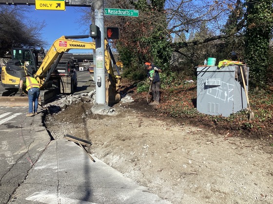 Crews demolishing northeast corner of Rainier Ave S and S Massachusetts to construct ADA curb ramps and install pedestrian signal push button.