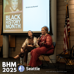 Two people sitting in front of a crowd by a screen that reads "Black History Month" 