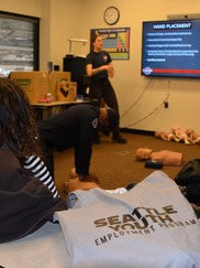 Seattle Youth Employment Program participants hear instructions from a White female EMT as a Black male fireman demonstrates on a CPR dummy