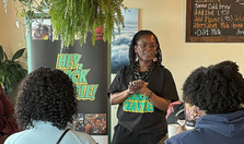 A Black women wearing a shirt that says "Hey! Black Seattle" speaks to a small group