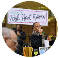 A Black woman speaking into a microphone with a sign hanging that reads: "Celebrating the High Point Mommas"
