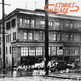 Black and white photo of a four story brick building at the corner of a city intersection with orange text: "Stories in Place"