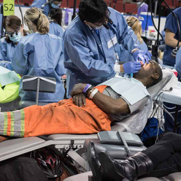 A person laying on a dental chair receiving an exam with many medical professionals and patients pictured in the background.
