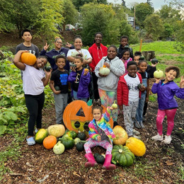An intergenerational group of mostly Black youth standing behind a pile of pumpkins smiling