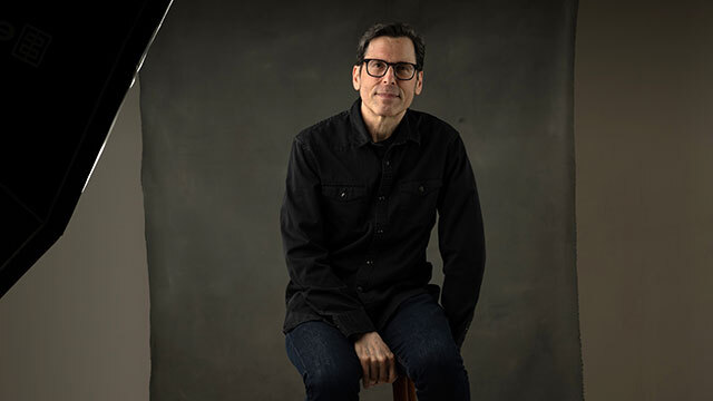 Man with dark hair, clothing, and glasses sits on wooden stool