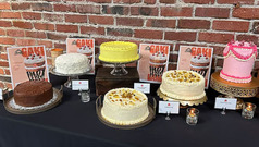 Six cakes with different color frosting displayed on a table in front of a brick wall