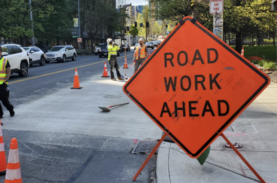 Image of 'Road Work Ahead' sign with cars, lane closures and construction workers on Denny Way in the background