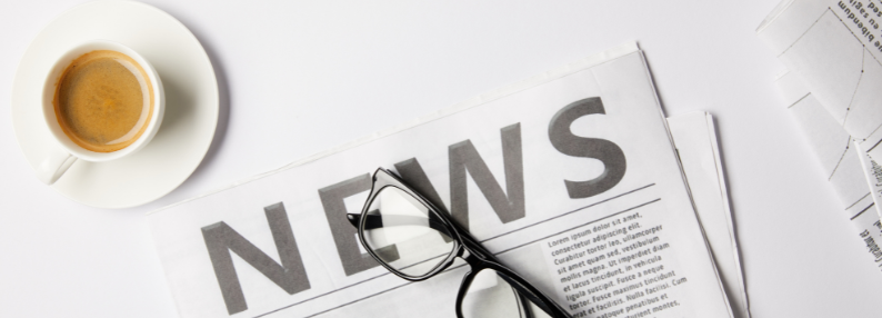 Tabletop with a coffee cup that has a latte in it, a newspaper that says 'News' at the top, and a pair of black reading glasses sitting on top of it.