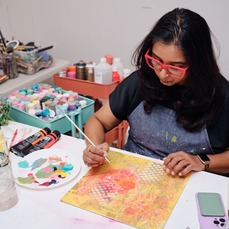 Woman with dark hair and pink framed glasses sitting at a desk working on a painting