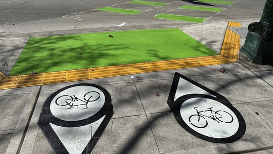 The green panel on the bike lane that rosses the intersection is wider, with two directional arrows to help cyclists know their position.