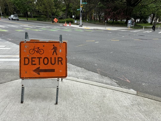 Image of 'bicycle and pedestrian detour' sign on sidewalk and vehicle traveling on Denny Way