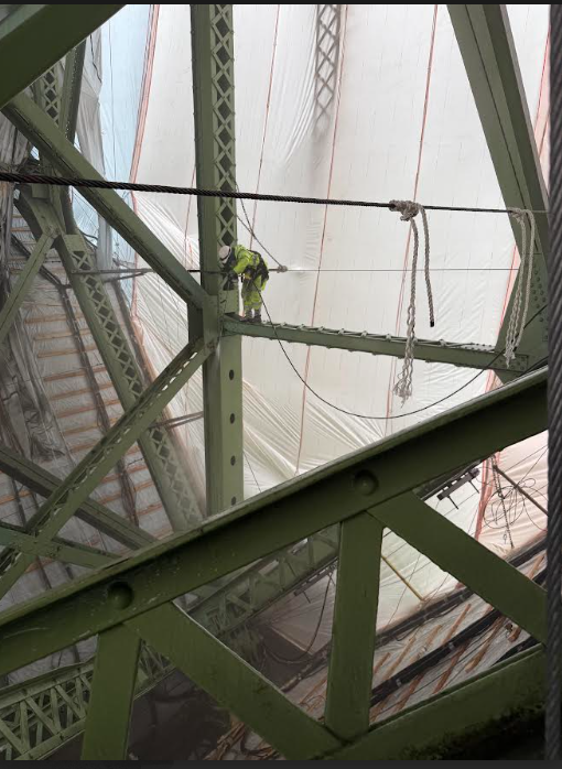 Worker on steel beams inside bridge frame, wearing safety gear, with scaffolding and containment sheeting visible.