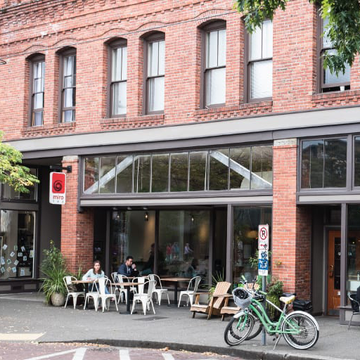 People seated at cafe tables on the sidewalk in front of a brick building with large storefront windows
