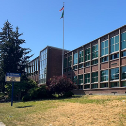 A two-story building with windows along both floors with blue message board and flag pole on grass in front