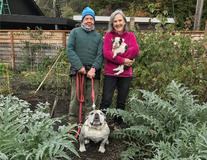 Two older woman standing in a garden plot, one holding a leash with a large bulldog and the other with a small dog in her arms
