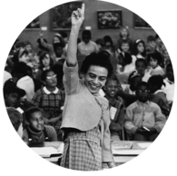 A black and white photo of a Black woman raising her hand in the air with one finger raised with a crowd pictured behind her
