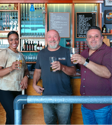 A woman and two men standing in front of a brewery bar, each holding a beer