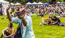 A person wearing a rainbow crown, dancing with a large crowd of people sitting on the grass in the background