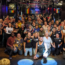 A group of 40-50 smiling people posing for a photo inside a bar with Pride flags hanging 