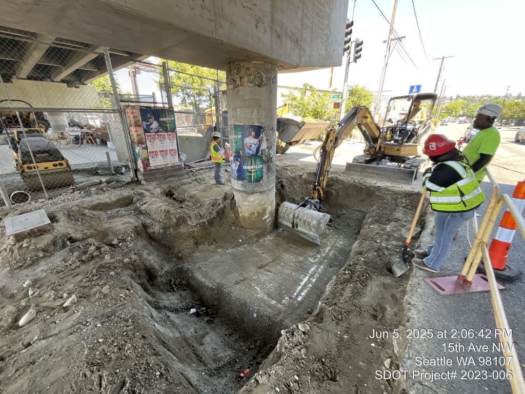 Crews demolish pavement and excavate soil to prepare for bridge footing retrofit work.
