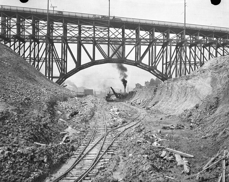Black and white image of steel bridge with railroad tracks, instead of street, underneath it.