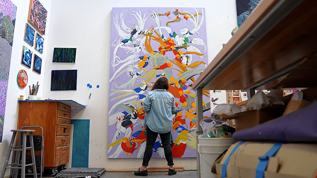 Artist with back to camera works on large purple painting with multicolored birds in art studio