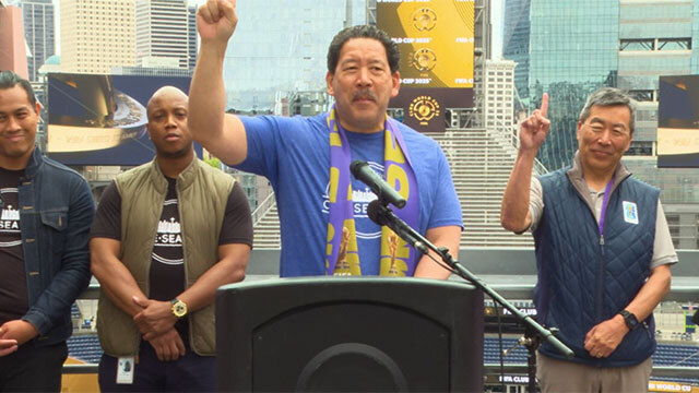 Mayor Bruce Harrell stands at podium with hand raised, three people stand behind