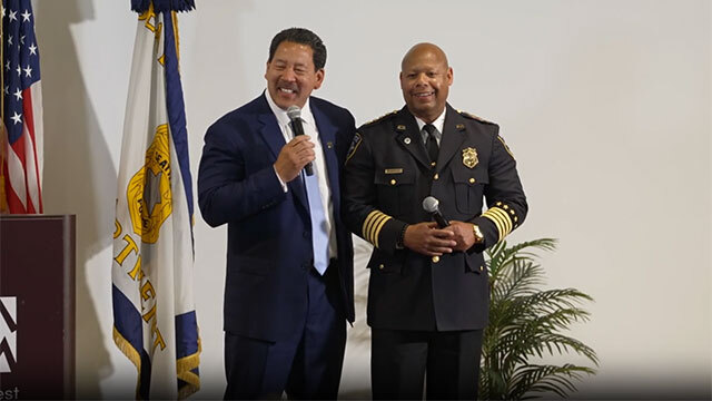 Seattle Mayor Bruce Harrell and Seattle Police Chief Barnes stand smiling in front of while wall and flags