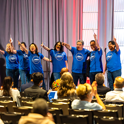 Seven people standing on a stage holding up their index fingers wearing blue shirts that say "One Seattle"