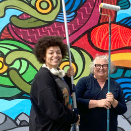 Two people standing in front of a colorful mural holding paint rollers