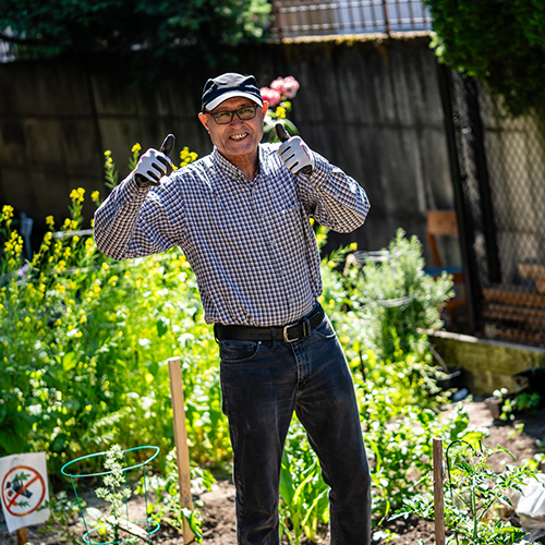A person wearing a hat, long sleeve shirt, and garden gloves standing in a garden giving two thumbs up
