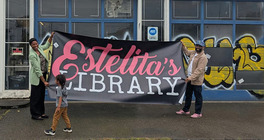 Two people standing in front of an industrial garage door holding a large sign that says "Estelita's Library" 