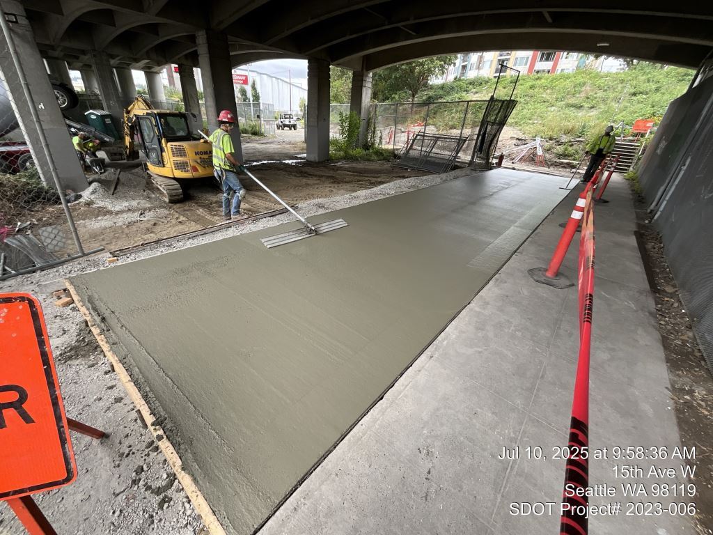 Crews remove existing fencing and pour a new, wider pedestrian walkway beneath the Ballard Bridge. Image taken July 10, 2025.