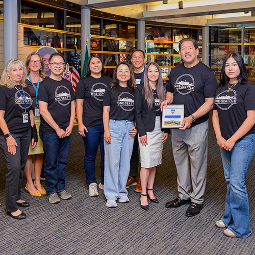 A group of people wearing black shirts that say "One Seattle" stand together with holding a plaque
