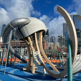 A playground with a jellyfish play structure in the foreground and tall city buildings pictured in the background