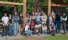 A large group of people, mostly youth, pose beneath a wooden structure outdoors