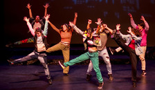 Seven people dancing on stage with one foot kicked out in the air and both hands raised high