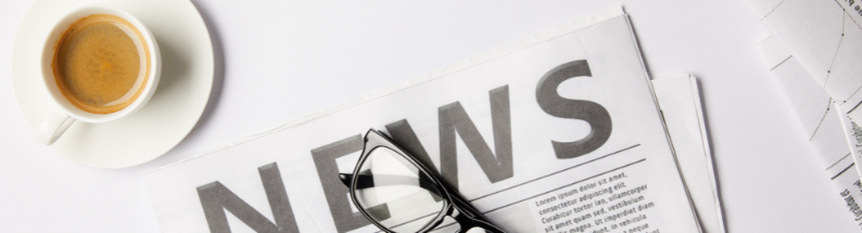 Photo of a newspaper on a white table, with folded up glasses sitting on top, and a coffee off to the side